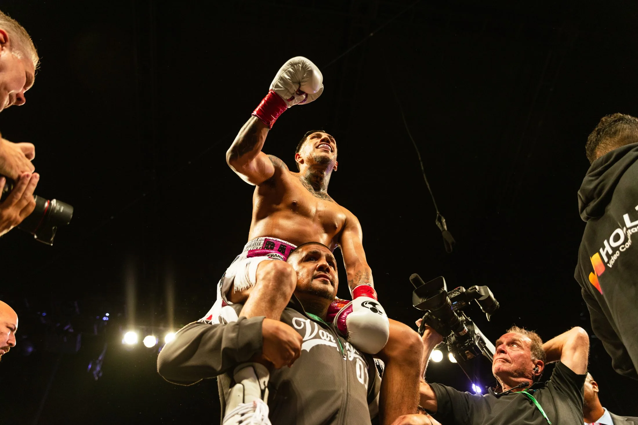 A fighter lifted on a cornerman's shoulders under arena light, glove raised, photographers and long lenses filling the press pit below